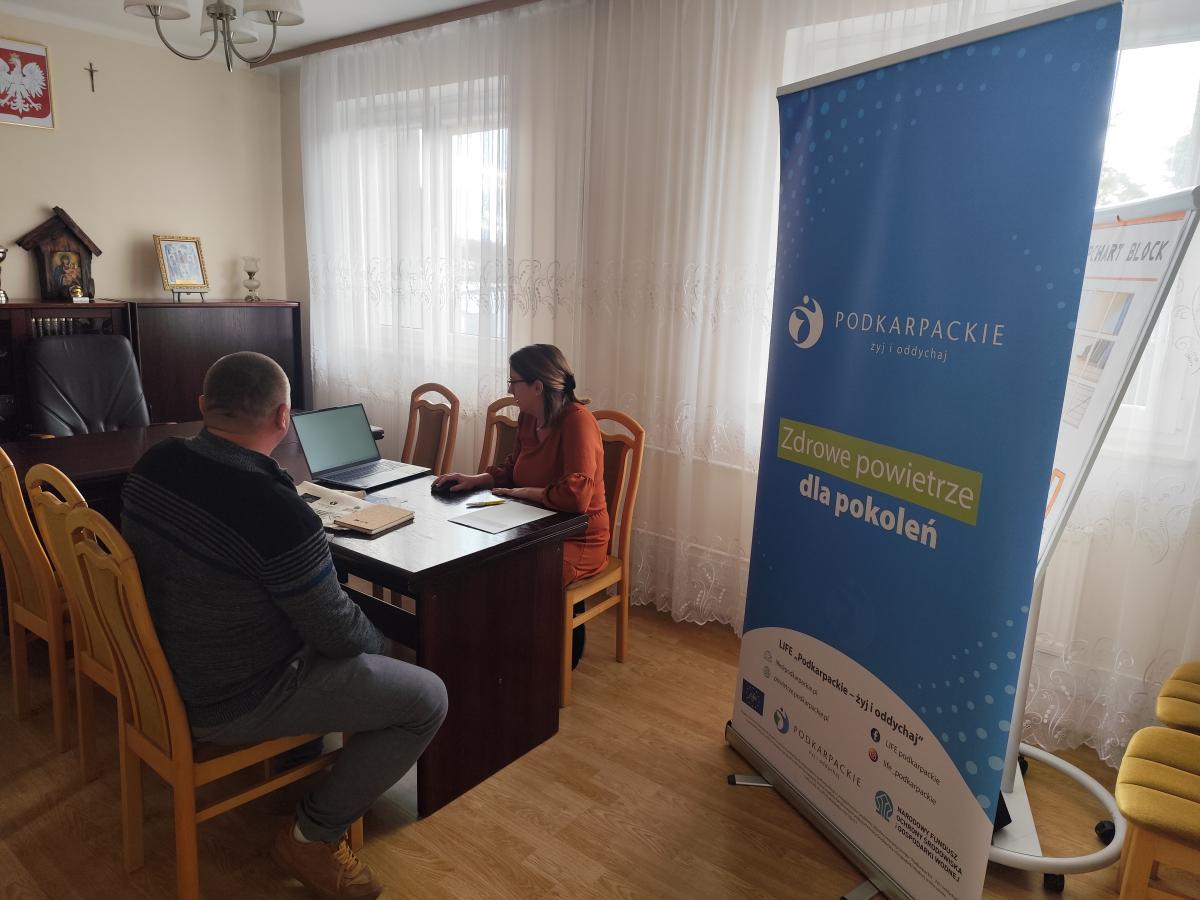 Wnętrze biura z osobami siedzącymi przy biurku i banerem z napisem "Zdrowe powietrze dla pokoleń" w tle. Na ścianie widoczny herb Polski.